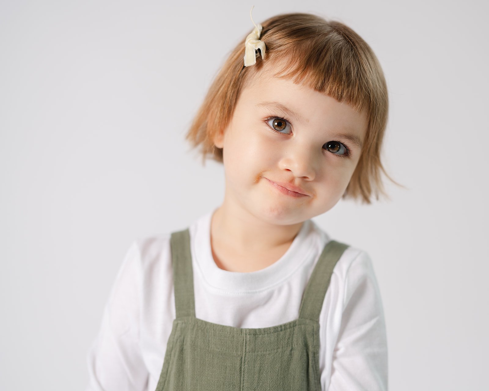 A young girl with short light brown hair, a hair clip, wearing a white shirt and green overalls, looking slightly to the side with a gentle smile.
