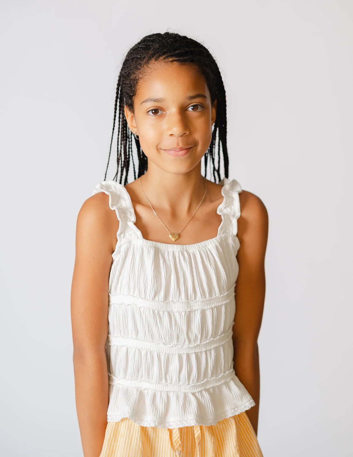 Young girl with braided hair wearing a white sleeveless top with ruffles and yellow striped shorts, standing against a plain white background.