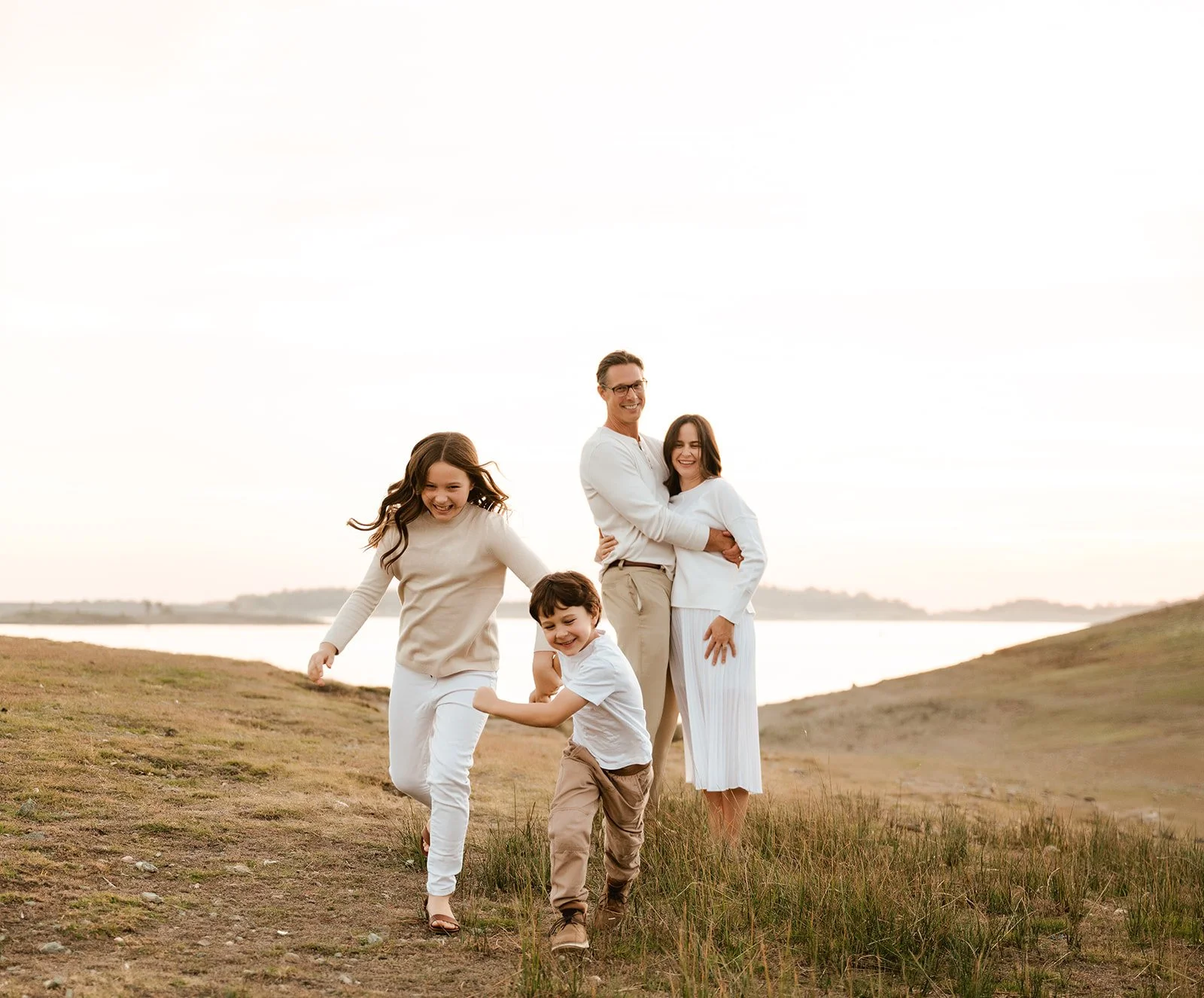 A happy family of five enjoying a day outdoors near a body of water during sunset. The family includes a father, mother, two daughters, and a son, all smiling and dressed in light-colored clothing.