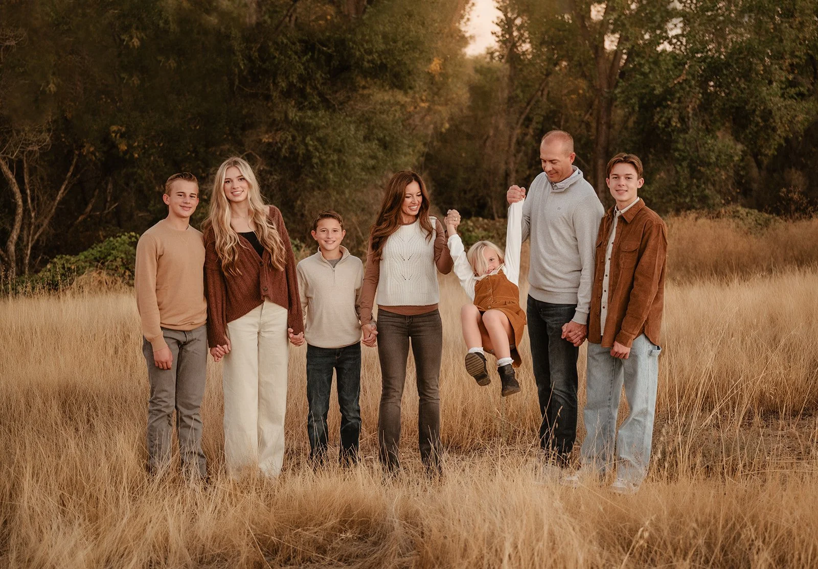 Family of eight standing outdoors in a field of tall, golden grass with trees in the background, holding hands, some smiling, and a young girl being lifted by her father.