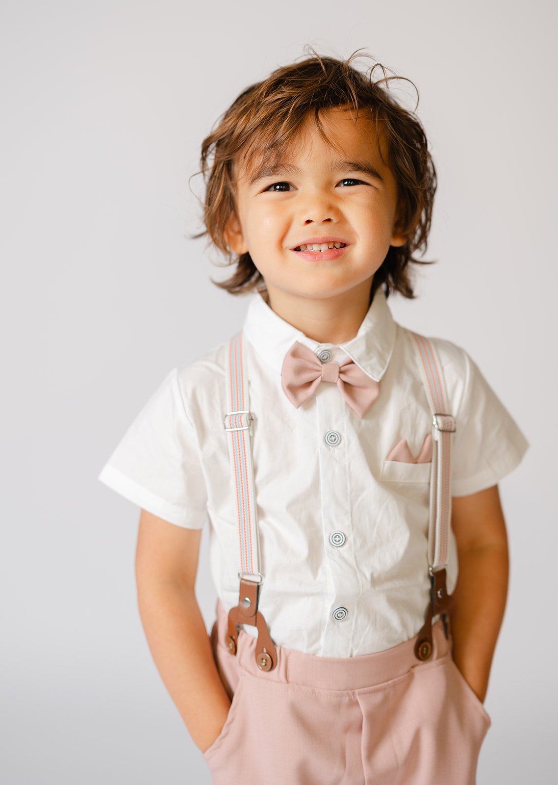 A young boy with curly brown hair smiling, wearing a white shirt with short sleeves, a pink bow tie, pink suspenders, and pink pants.