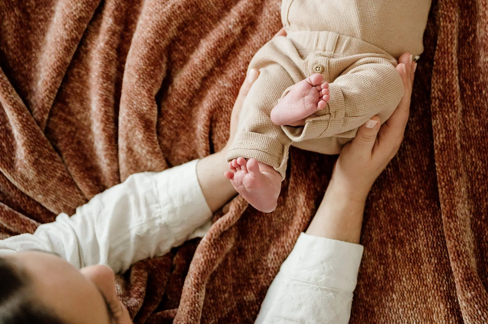 A tender moment showing a person cradling a baby on a textured brown blanket, focusing on the baby's tiny feet and relaxed limbs.
