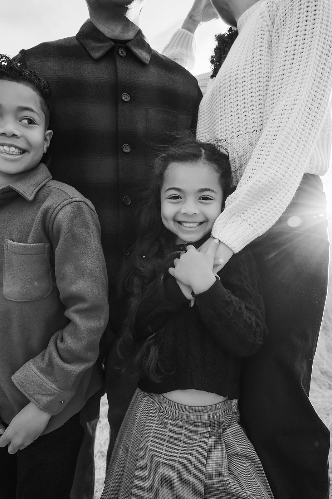 A black and white photo of a smiling young girl with long hair, standing with her hands clasped, surrounded by a boy and two adults, outdoors with sunlight.