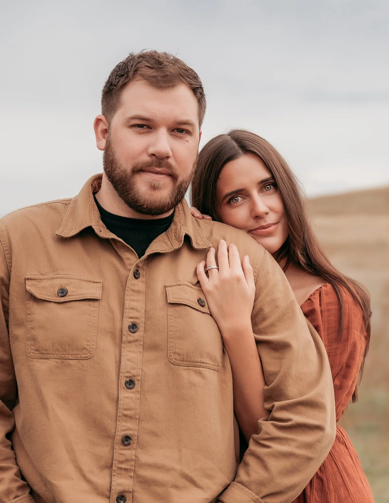 A man with short brown hair and a beard wearing a tan shirt, standing outdoors with a woman with long brown hair and blue eyes, wearing a rust-colored dress. They are close together, with the woman resting her hand on the man's shoulder, displaying a