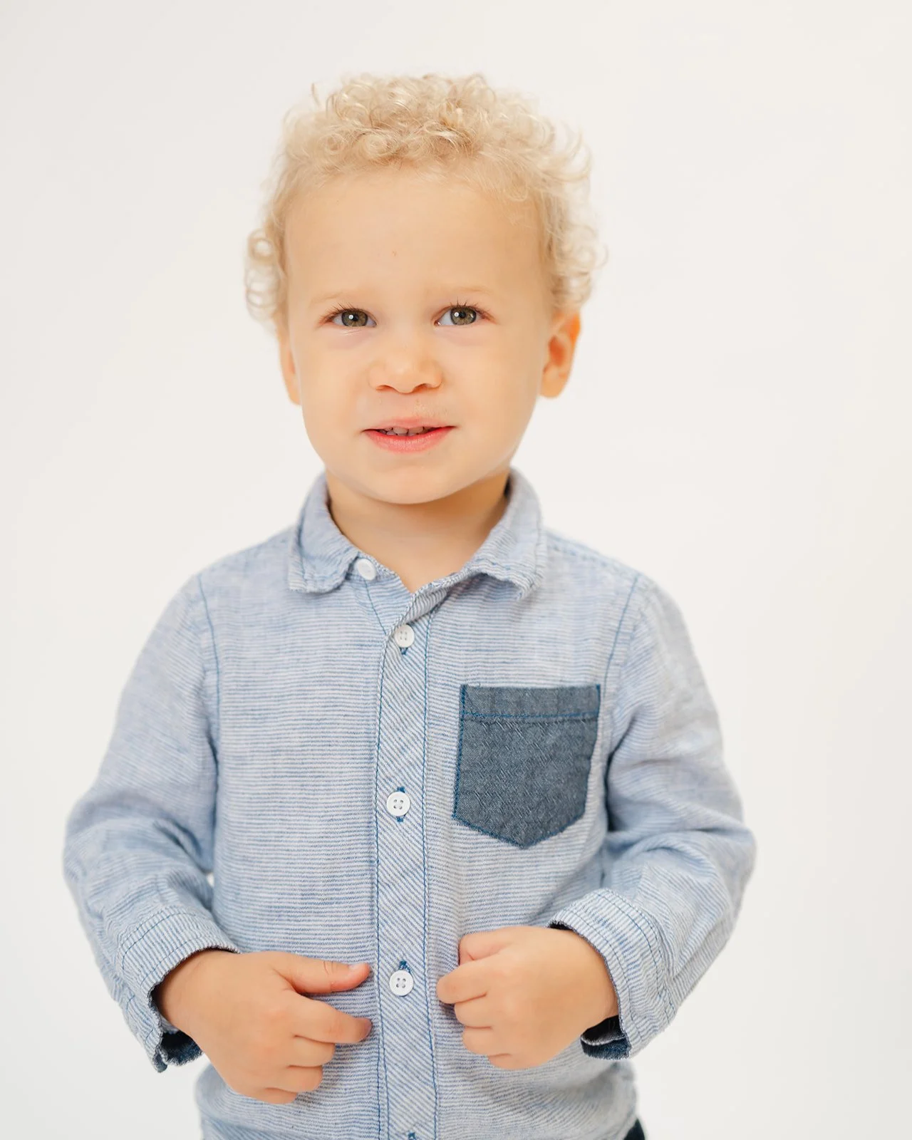 A young blonde boy with curly hair and blue eyes wearing a light blue button-up shirt with a dark blue pocket, standing against a plain white background.