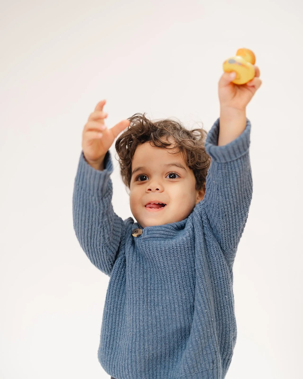 Child in a blue sweater raising arms and holding a yellow toy, with a white background.
