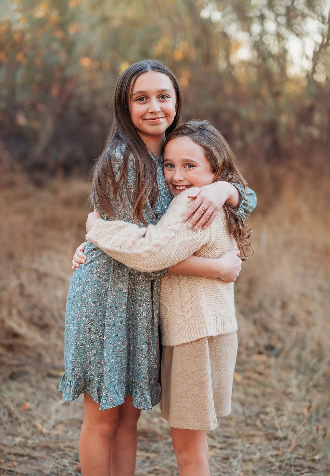 Two young girls hugging outdoors during sunset. The girl on the left has long brown hair and is wearing a blue patterned dress. The girl on the right has wavy brown hair and is wearing a cream-colored sweater and shorts. They are smiling and standing