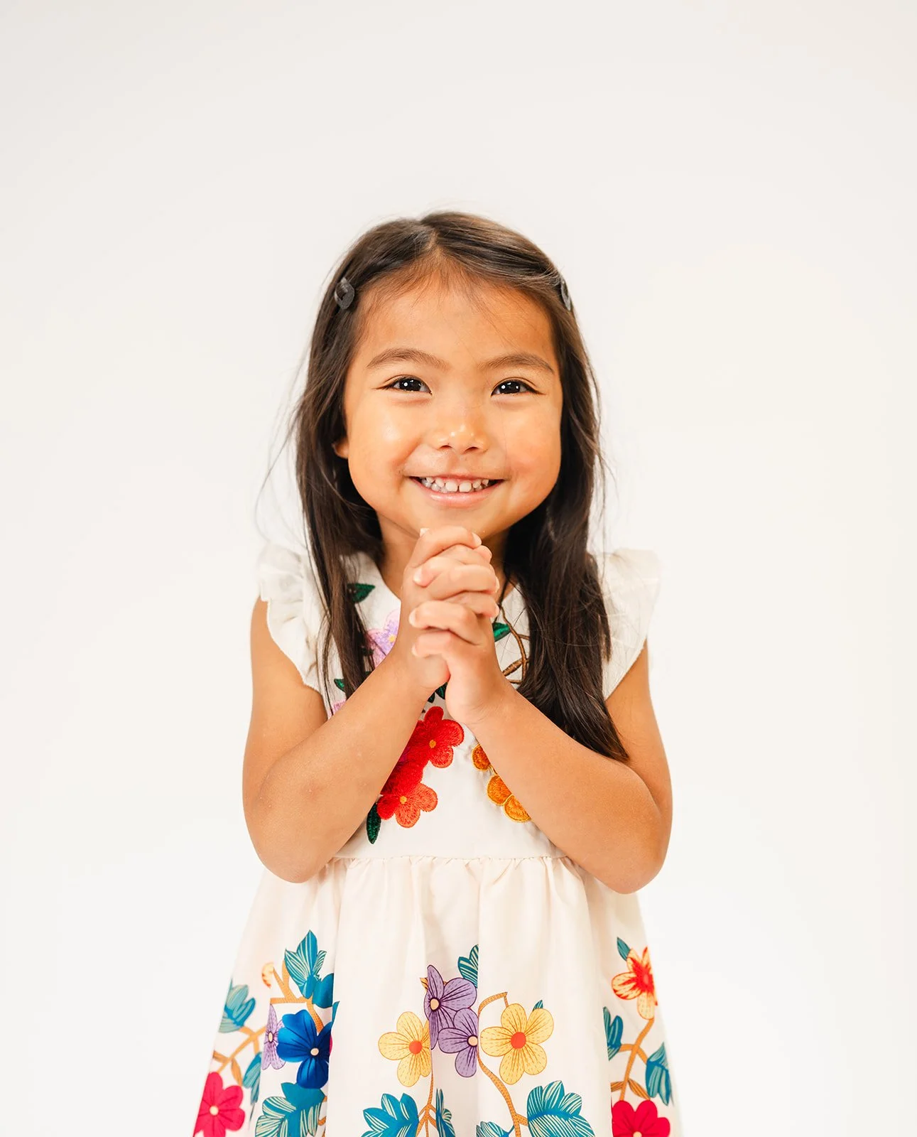 Smiling young girl with long dark hair, wearing a white dress with colorful floral embroidery, clasping her hands together.