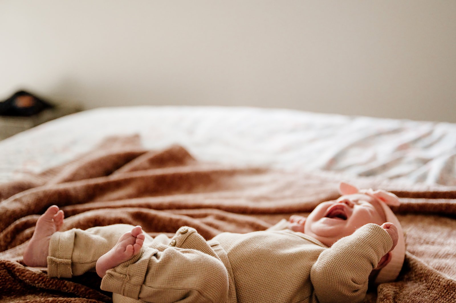 A baby lying on a bed, laughing and wearing a beige onesie and a pink headband with a bow, on a brown blanket.
