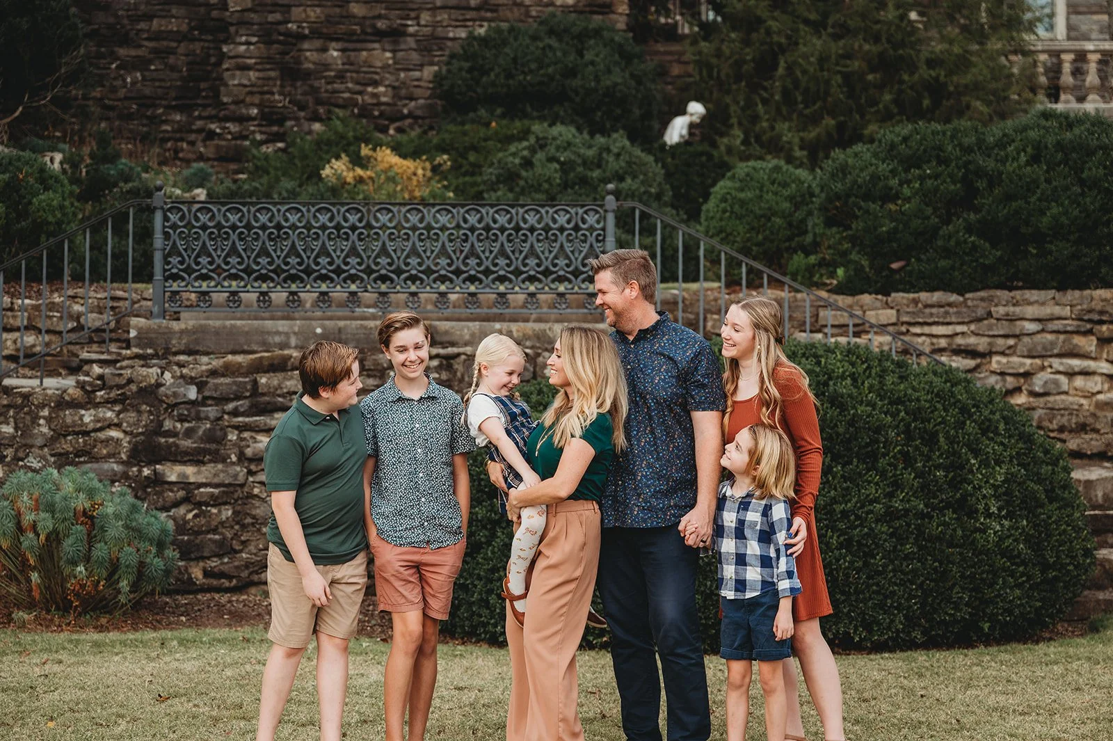 Family of seven standing outdoors on grass, laughing and holding hands, with trees and stone steps in the background.
