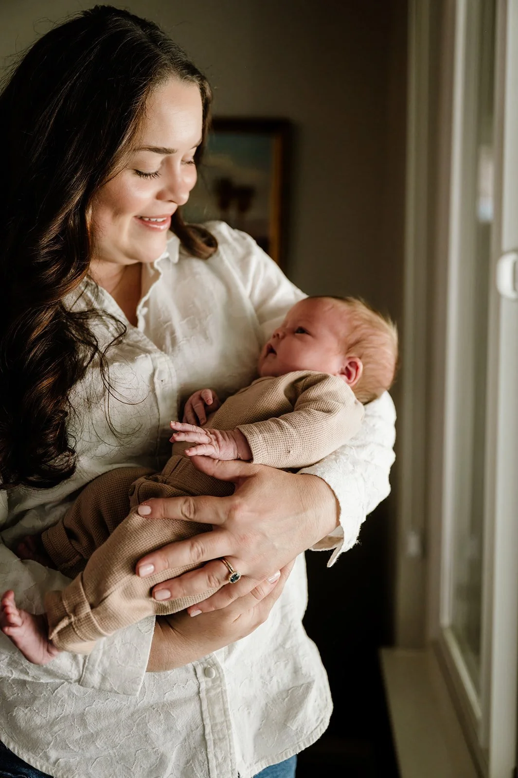 A woman holding a newborn baby near a window, both looking at each other lovingly.