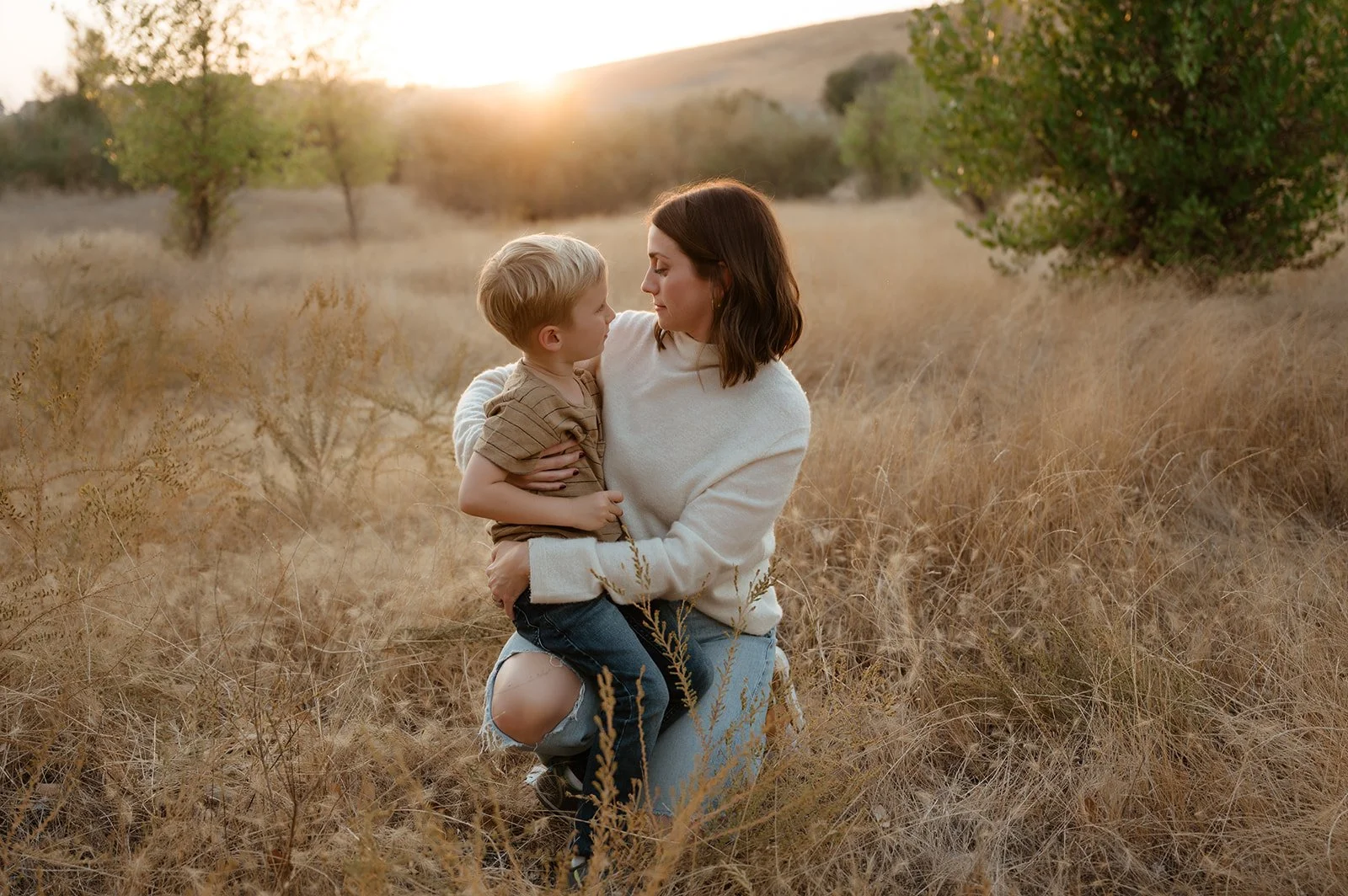 A woman holding a young boy in a field of tall, dry grass during sunset.