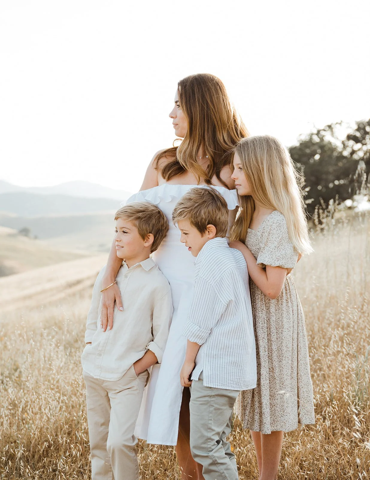 A woman and three children standing in a field with tall grass, holding each other, during sunset.
