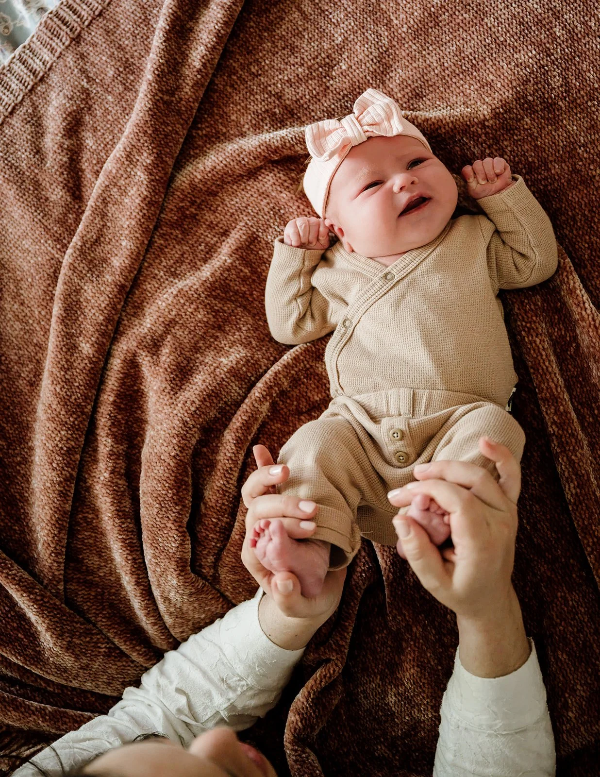 A baby lying on a brown textured blanket, wearing a beige outfit and a white headband with a pink bow, smiling with arms raised.