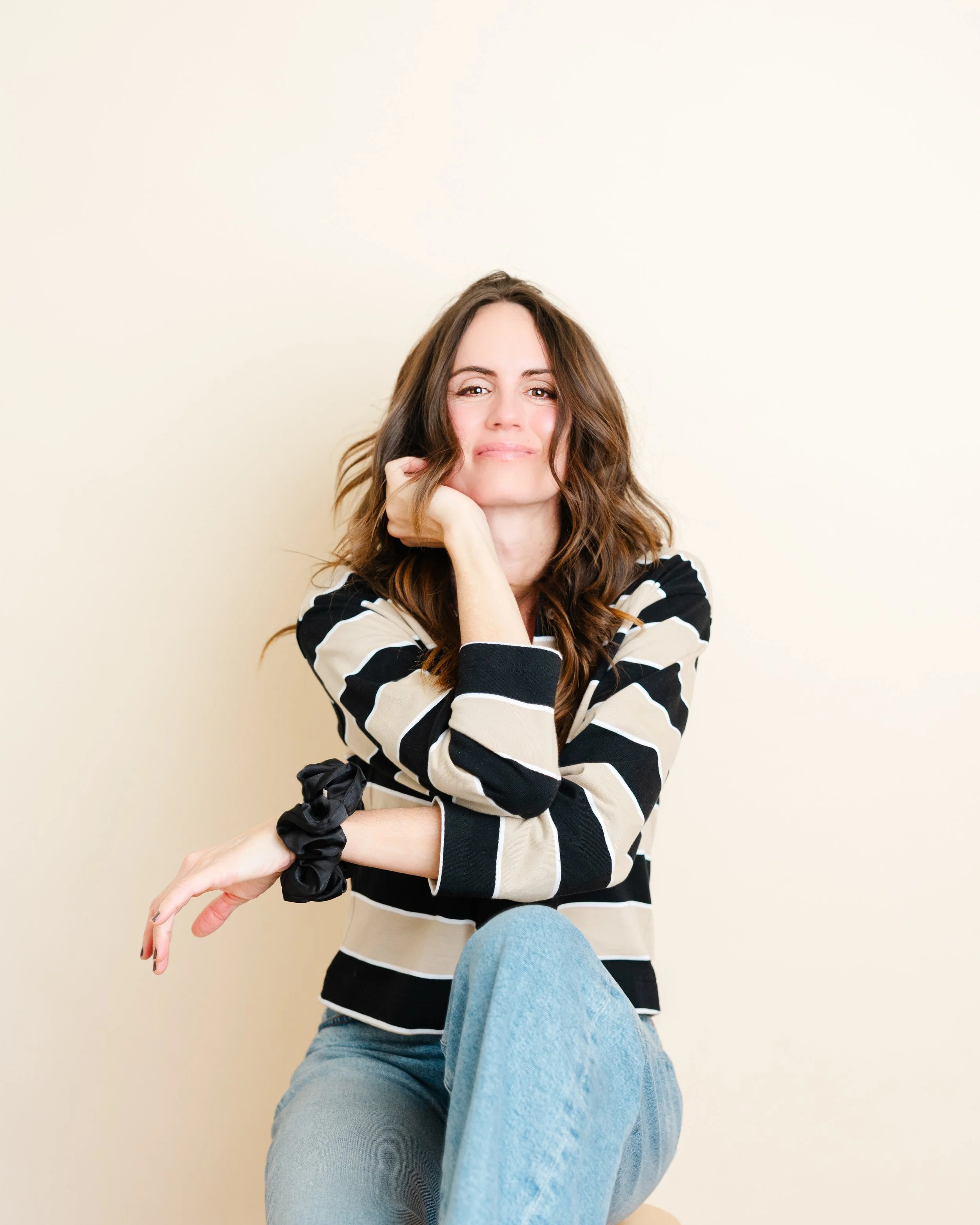 A woman with long, wavy brown hair and light skin, sitting with one knee up, wearing a black and beige striped long sleeve shirt and light blue jeans, posing with her chin resting on her right hand and looking at the camera with a slight smile, against a plain beige background.
