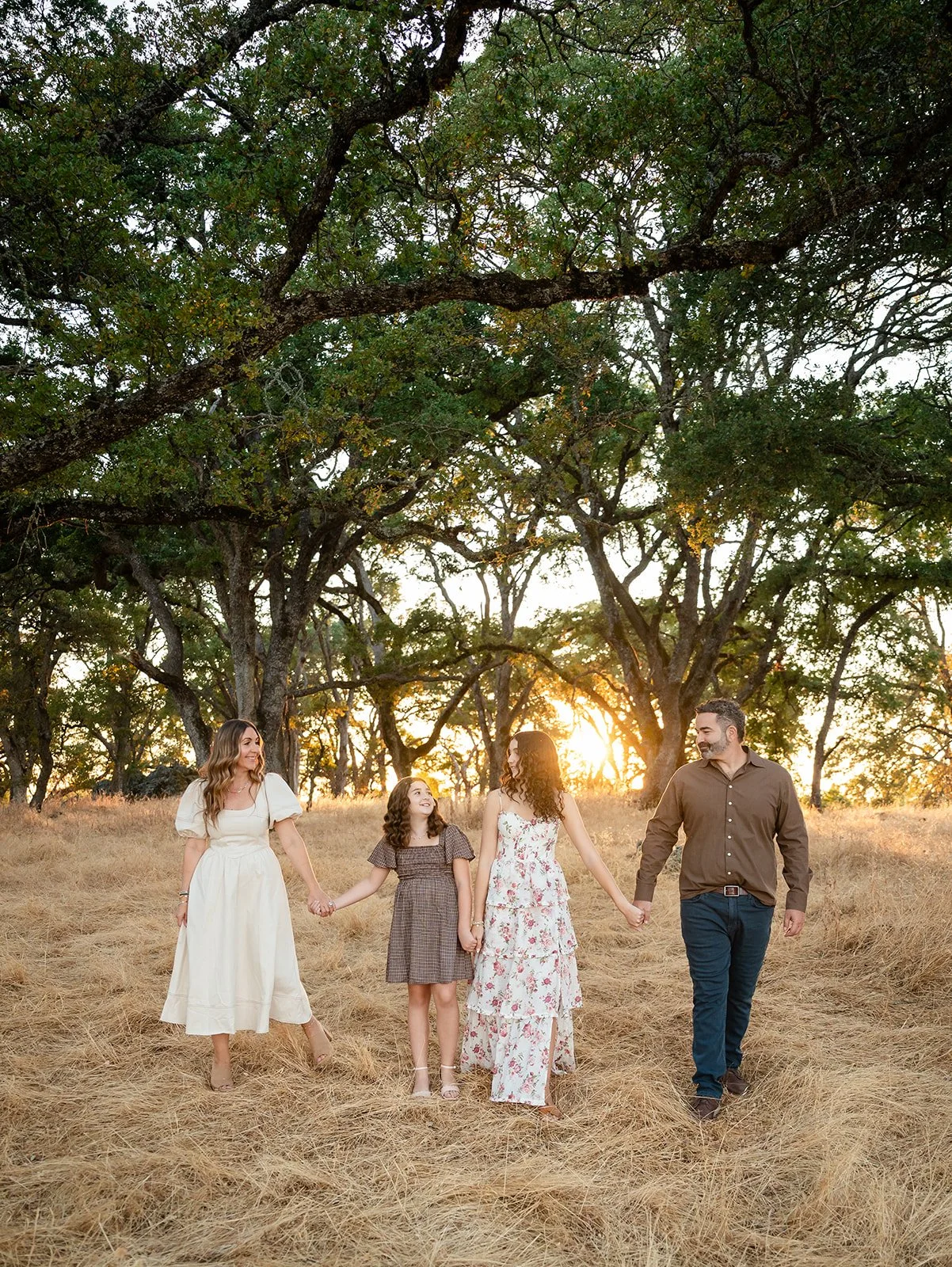 Family of four holding hands and walking in a field with trees in the background during sunset.