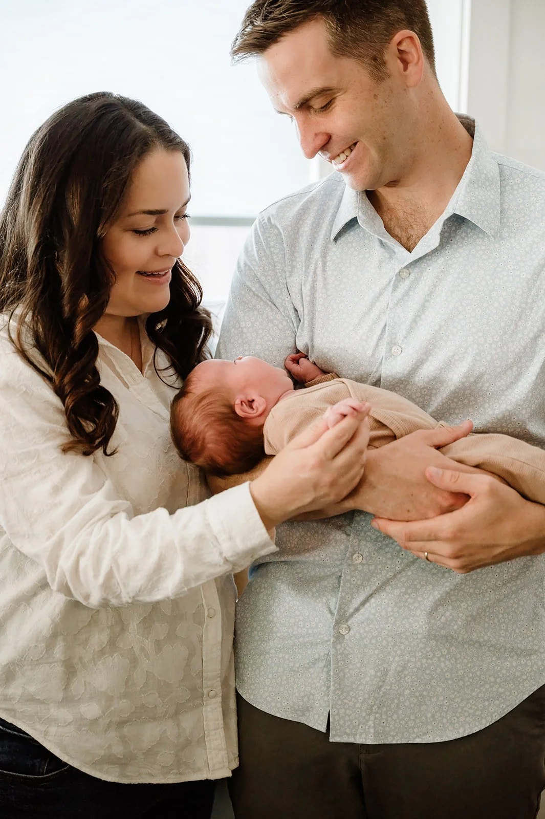 A smiling family, a woman and a man, are holding a newborn baby together in a brightly lit room.