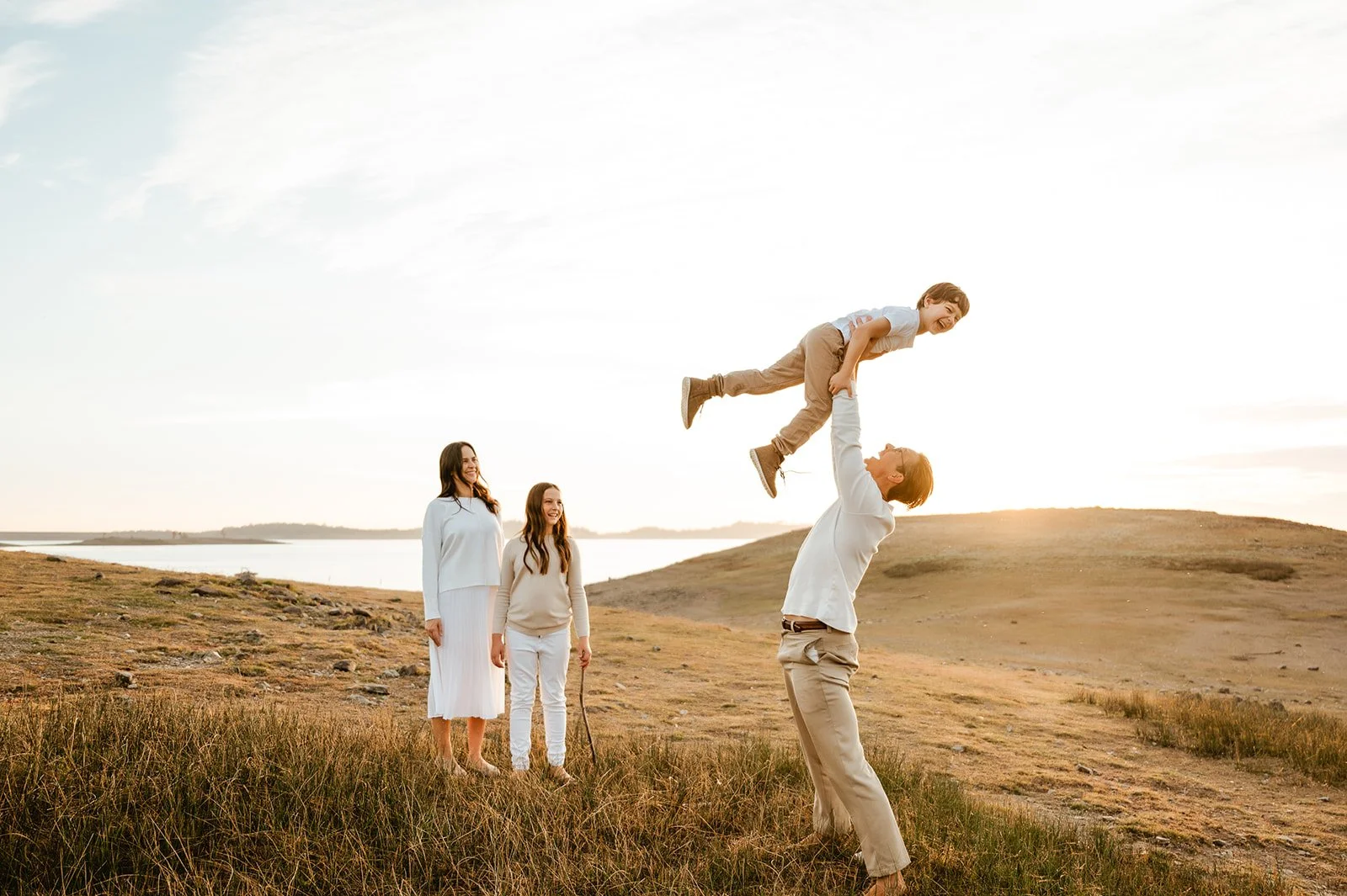 A family of four enjoying an outdoor moment at sunset, with a man lifting a young boy in the air, and a woman and girl watching and smiling.