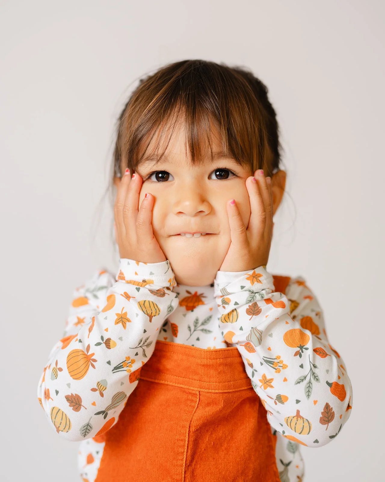 A young girl with brown hair and brown eyes, pressing her cheeks with her hands, wearing a white long-sleeve shirt with orange and green leaf and pumpkin patterns, and an orange overall.