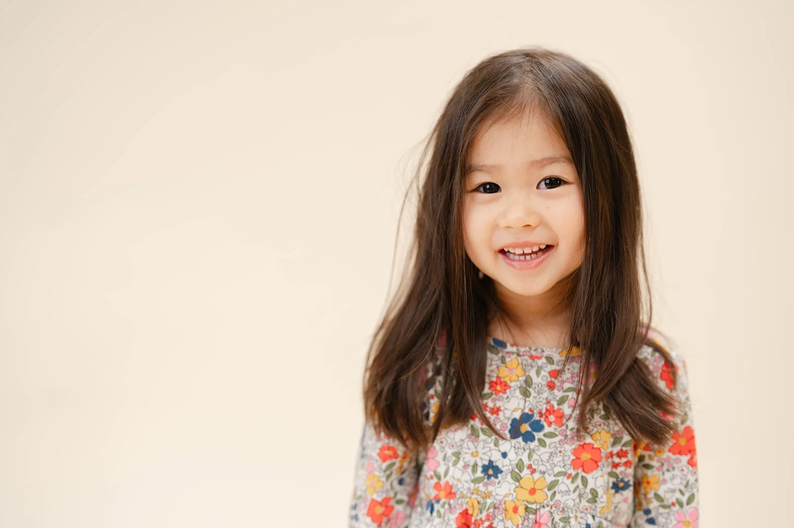 A young girl with long brown hair, smiling, wearing a colorful floral dress, against a plain beige background.