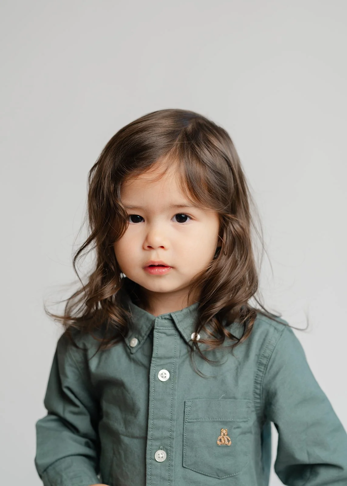 A young girl with long brown hair and dark eyes, wearing a green button-up shirt with a small emblem on the pocket, standing against a plain light gray background.