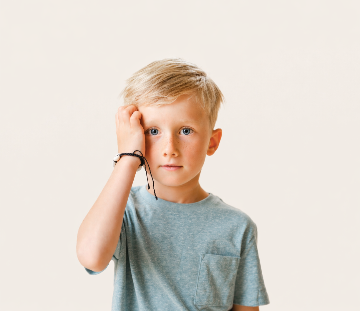 A young boy with blonde hair and blue eyes, wearing a grey t-shirt, touching his forehead with his right hand, standing against a plain background.