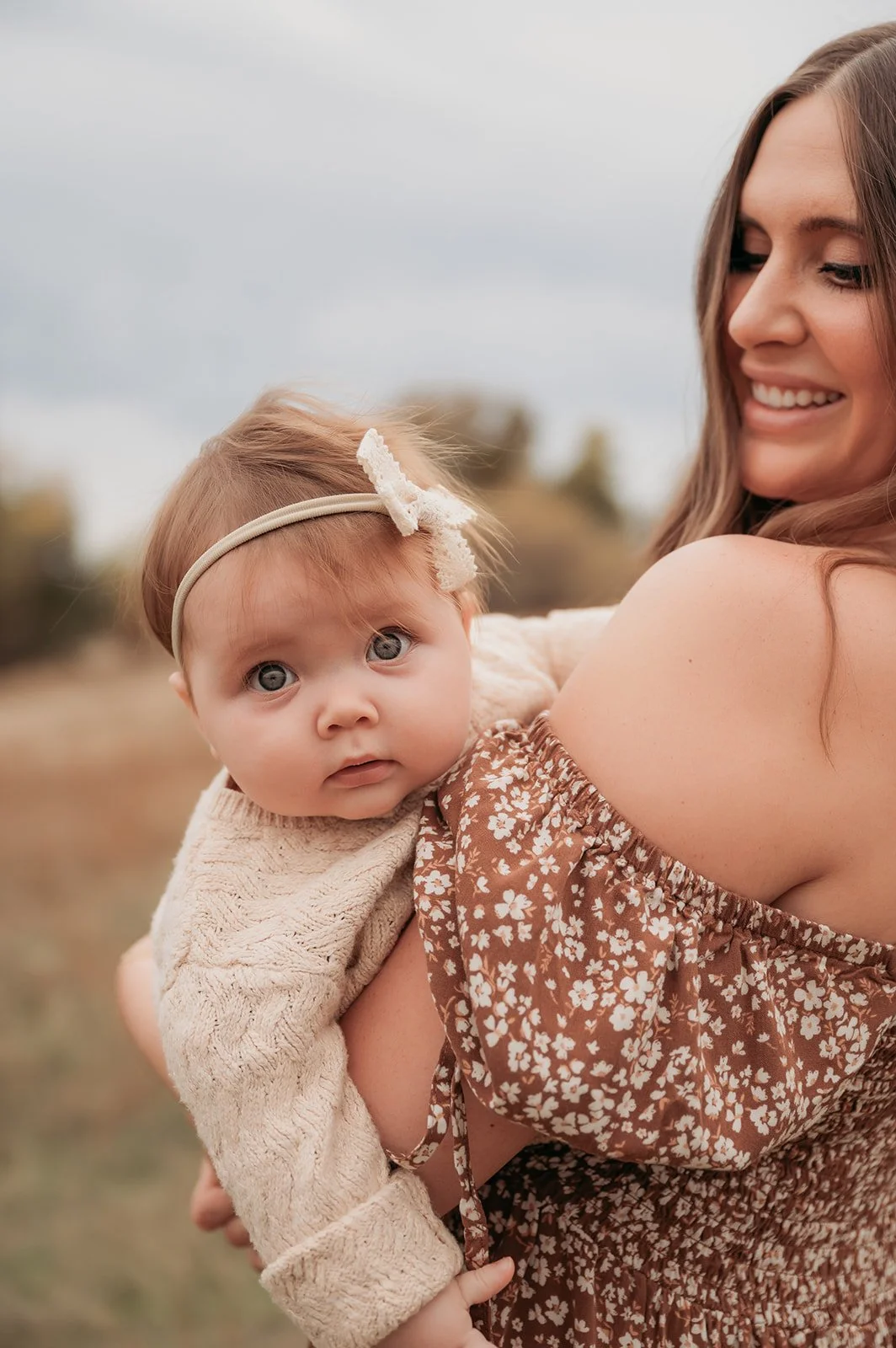 A woman holding a baby girl outdoors, both looking at the camera. The woman is smiling and wearing a floral dress, while the baby has a headband with a bow and a beige sweater.