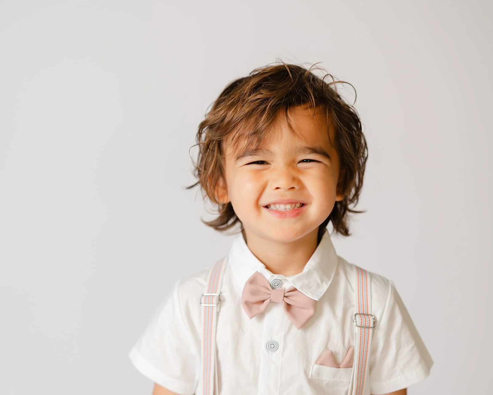 A smiling young boy with messy brown hair, wearing a white shirt with a pink bow tie and suspenders.