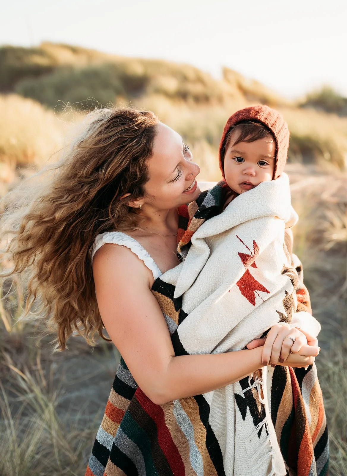 A woman holding a young child wrapped in a blanket outdoors with grassy dunes in the background.