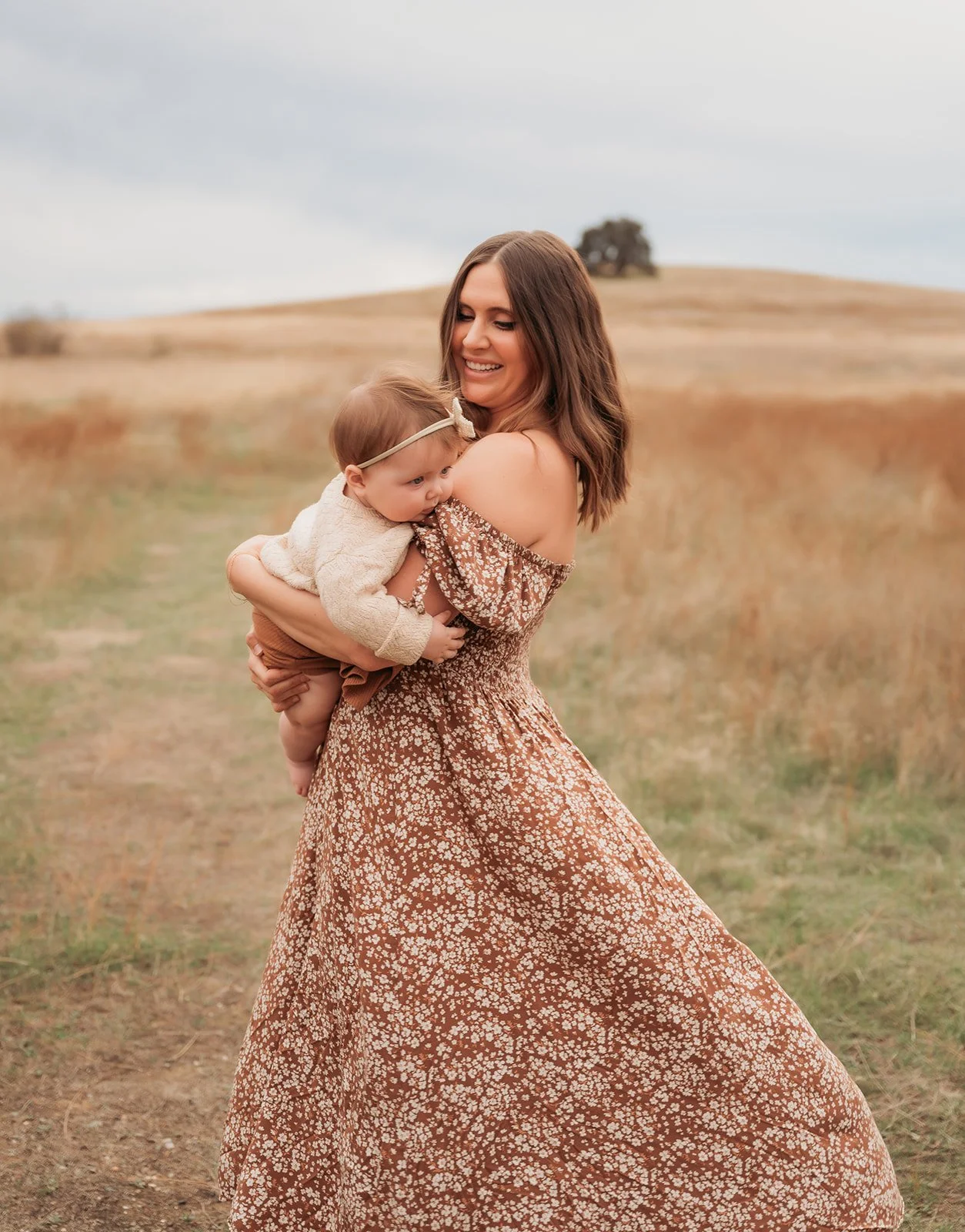 A woman in a flowy, floral dress holding a young child in an open field with a hill and a tree in the background.