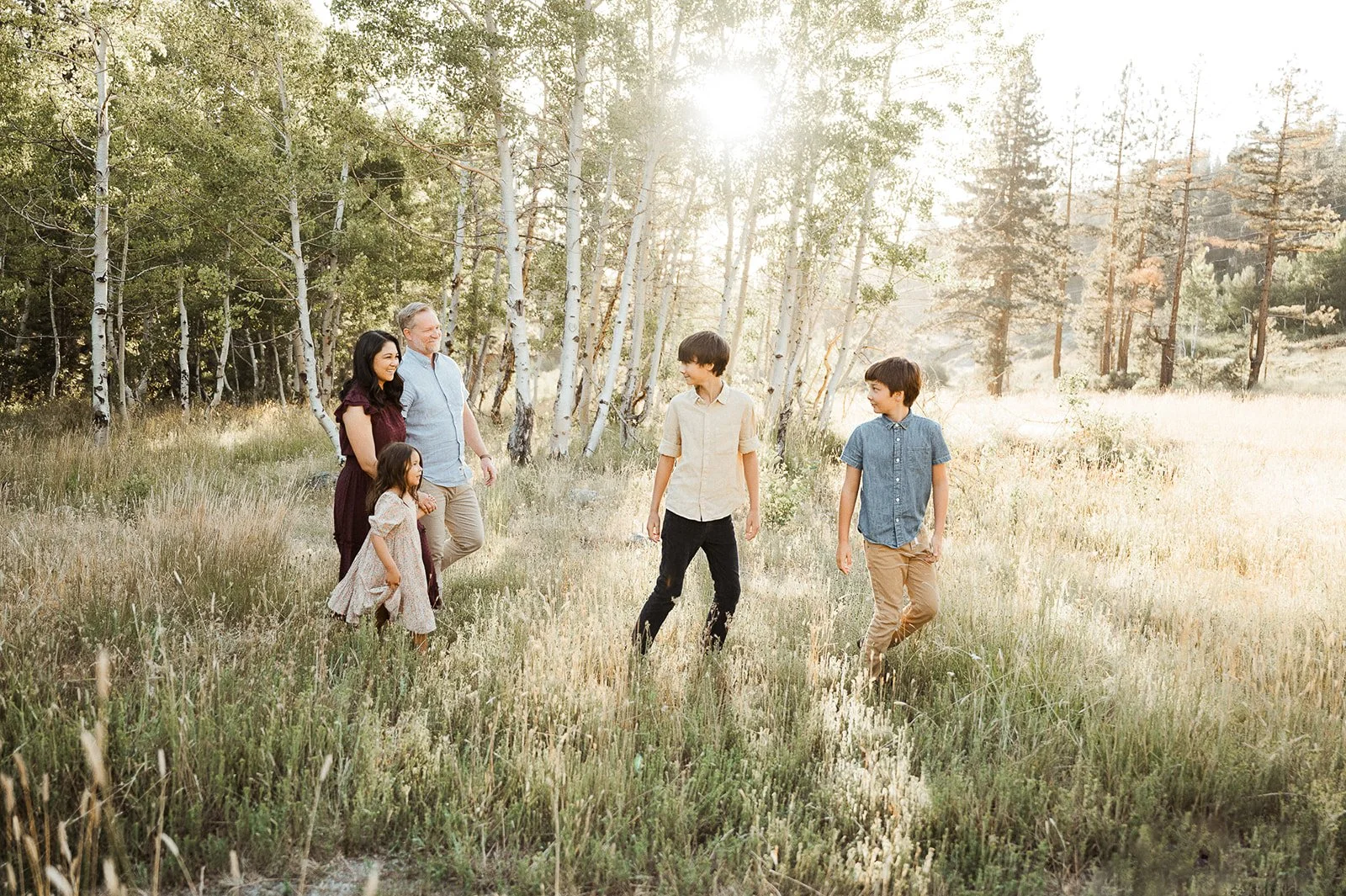 A family of five, including a man, woman, two boys, and a girl, walking through a grassy field with trees in the background during sunset.
