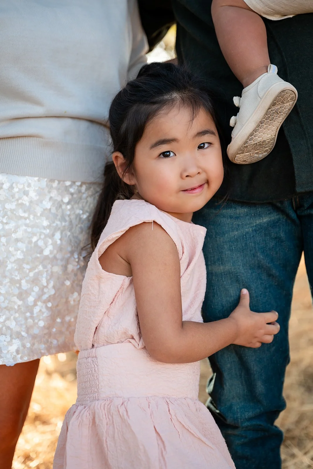 A young girl with long dark hair, wearing a light pink dress, is looking at the camera while leaning against an adult in dark jeans. The girl has a slight smile and her hand rests on the adult's leg. The background suggests an outdoor setting.