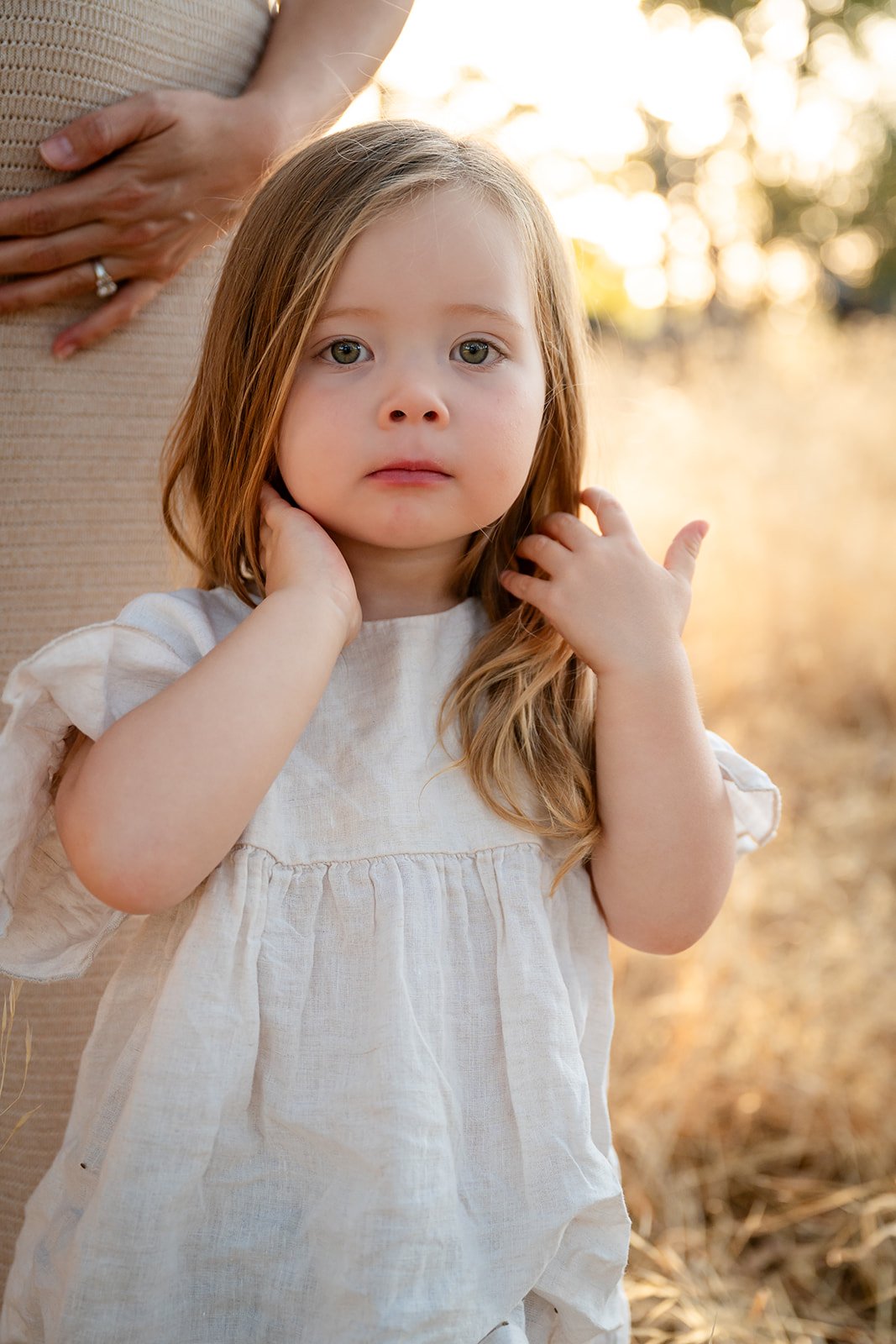A young girl with red hair and green eyes poses outdoors during sunset, with an adult partially visible in the background, wearing a light-colored dress, touching her neck and hair.