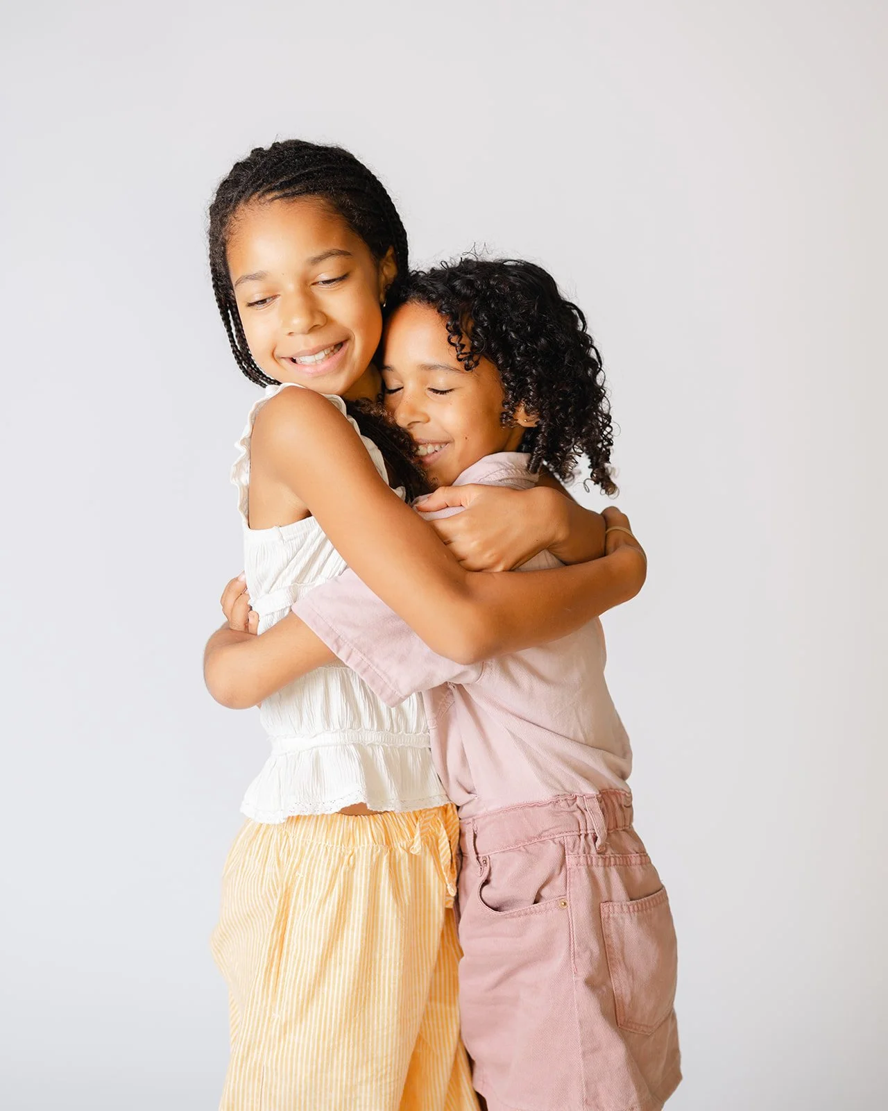 Two young girls hugging and smiling against a plain background.