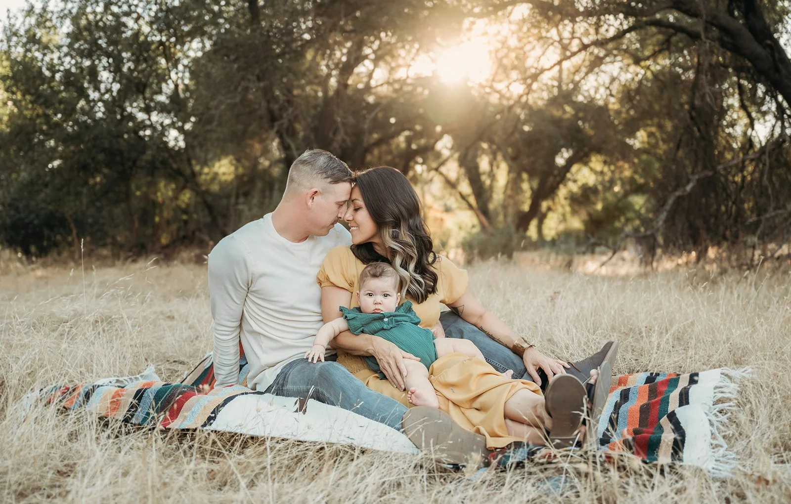 A family of three sitting on a blanket outdoors in a dry grassy area with trees in the background and sunlight shining through the trees.