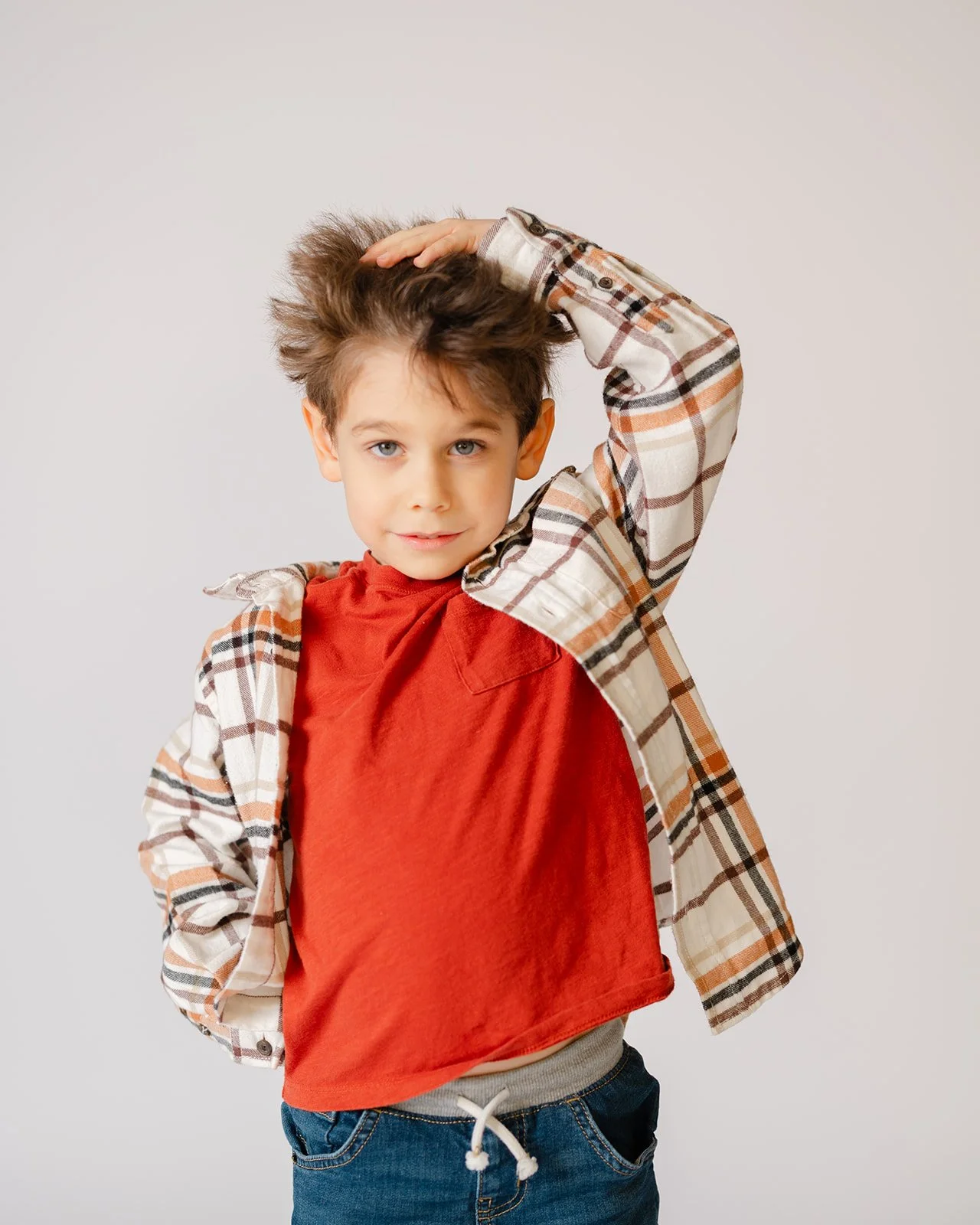 Young boy with tousled brown hair wearing a red t-shirt, plaid shirt, and jeans, standing against a plain light background, looking at the camera with a confident expression.