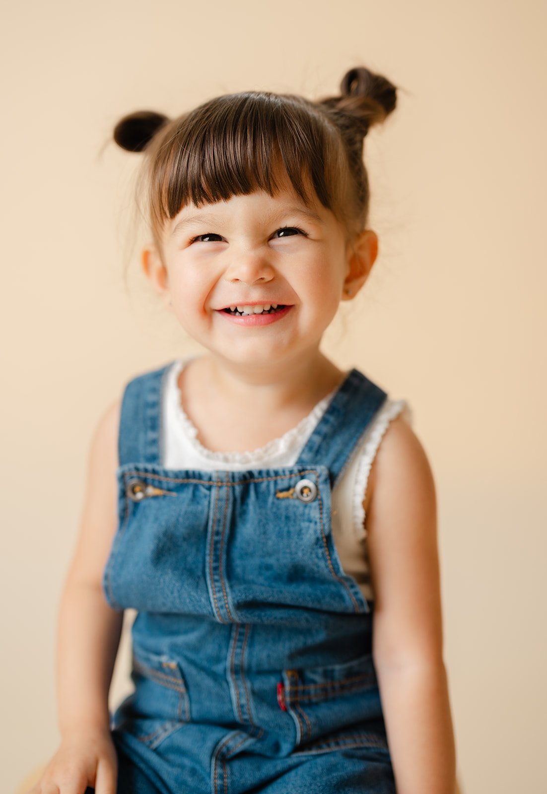 A young girl smiling with a gap-toothed grin, wearing denim overalls and a sleeveless white top, with her hair in two buns.