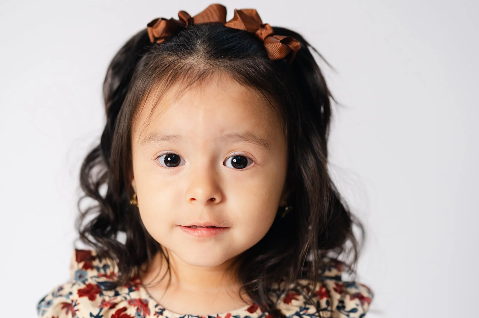 A young girl with dark curly hair wearing a floral dress and a brown bow headband, looking directly at the camera.