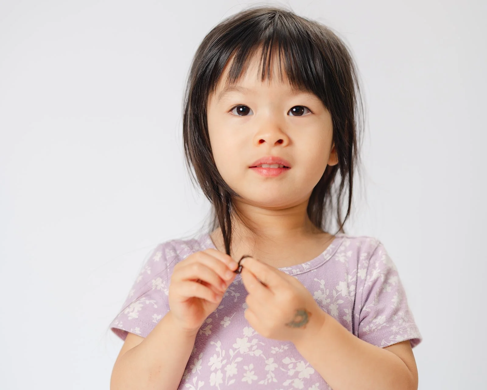 A young girl with medium-length dark hair and a floral pink shirt looking at the camera, holding her hands together.
