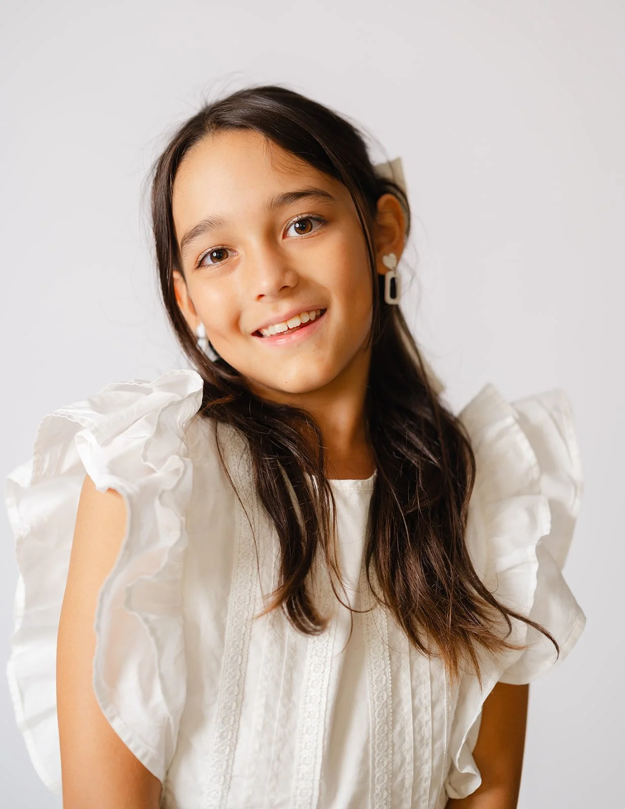 A young girl with long dark hair, wearing a white ruffled dress and earrings, smiling at the camera against a plain light background.