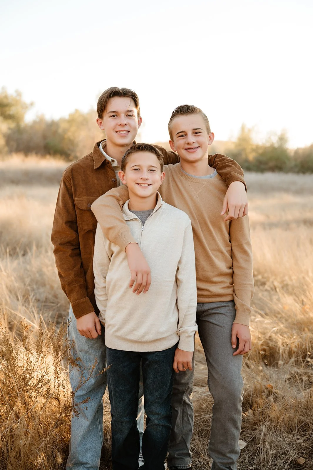 Three young boys standing outdoors in a field, smiling, with one boy having his arms around the other two, during sunset with soft natural lighting.