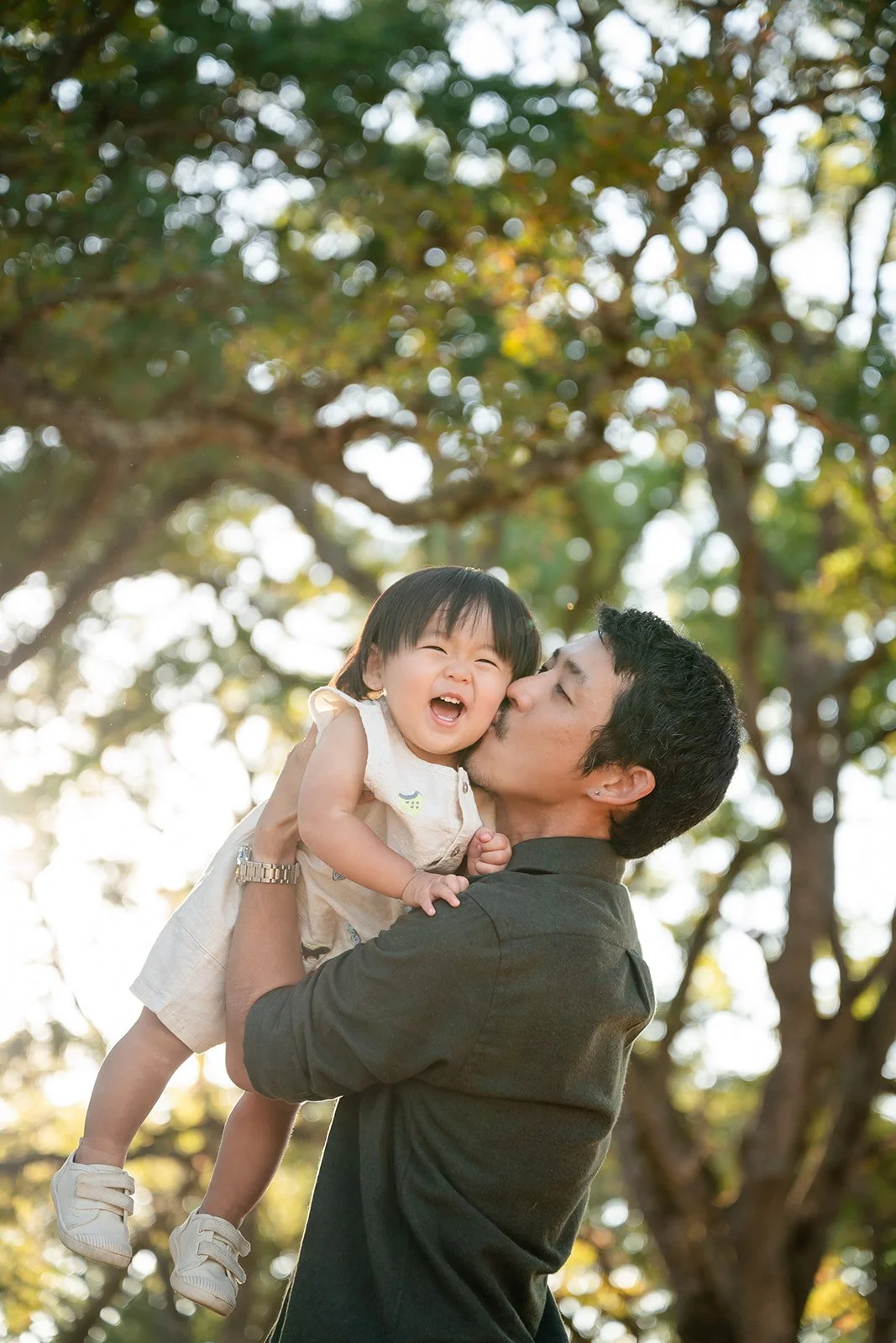 A man holding a laughing young girl in an outdoor setting with trees and sunlight.