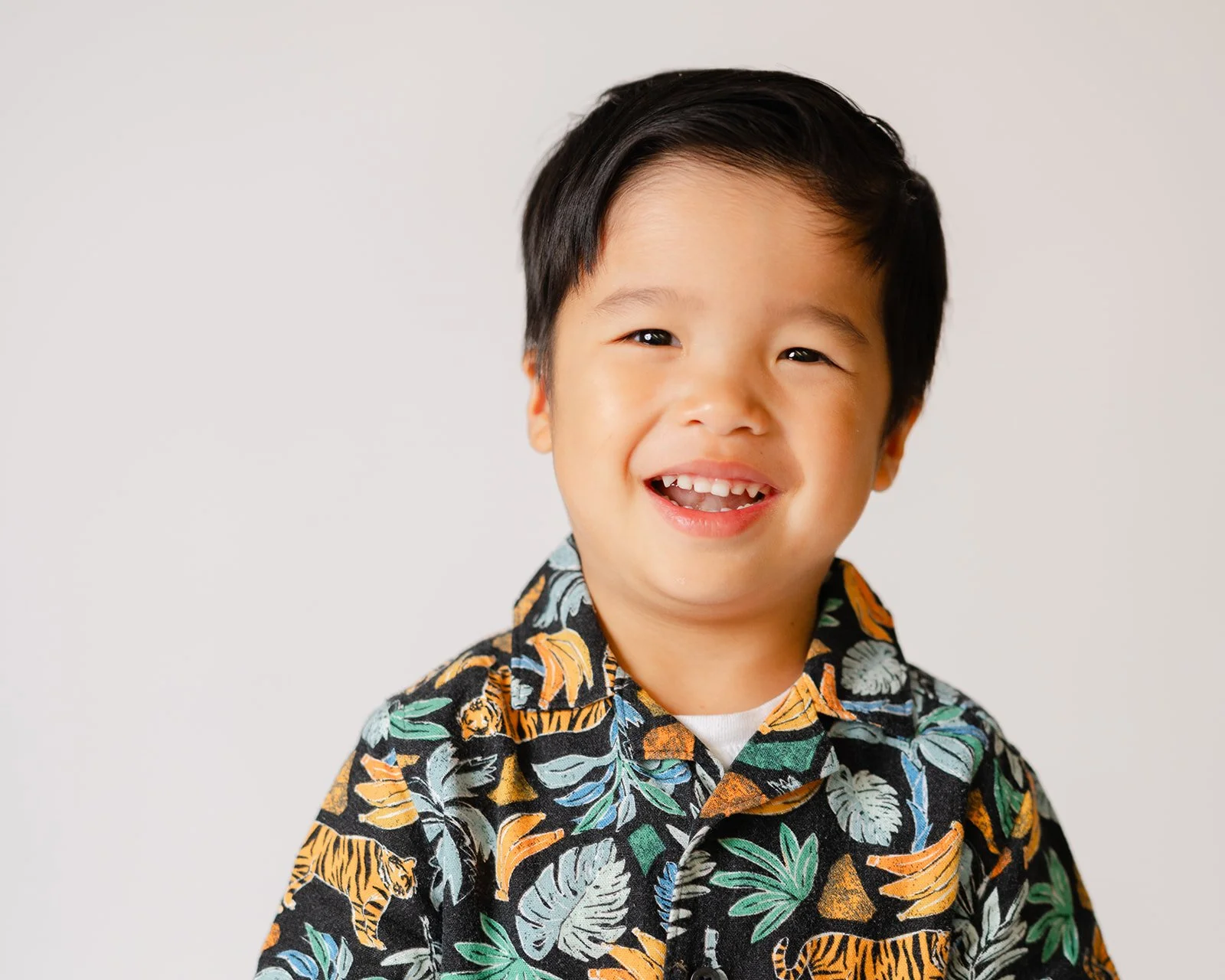A young boy with black hair, smiling and showing his teeth, wearing a colorful short-sleeved shirt with a jungle theme, against a plain white background.