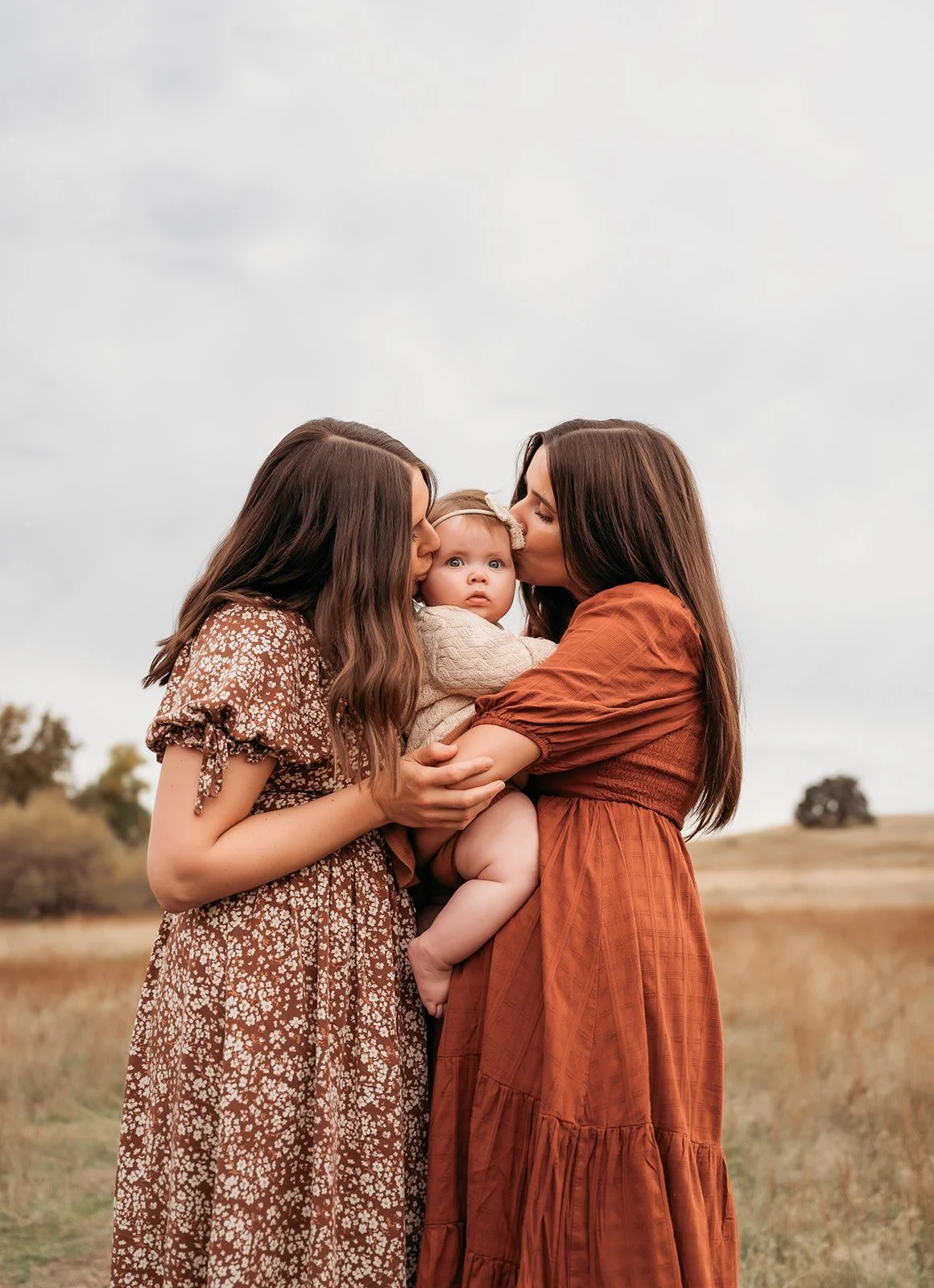 Two women kissing a baby girl on the cheeks in an outdoor field with cloudy sky and trees in the background.