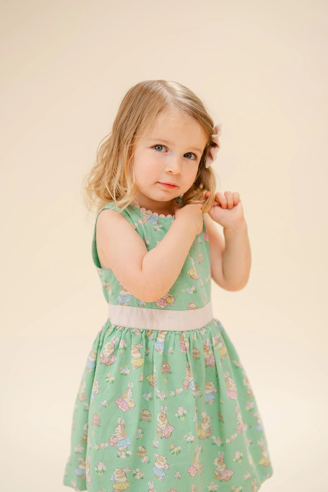 A young girl with blonde hair and blue eyes, wearing a sleeveless green dress with pastel bunny and floral patterns, standing against a plain beige background with her hands clasped near her shoulders.