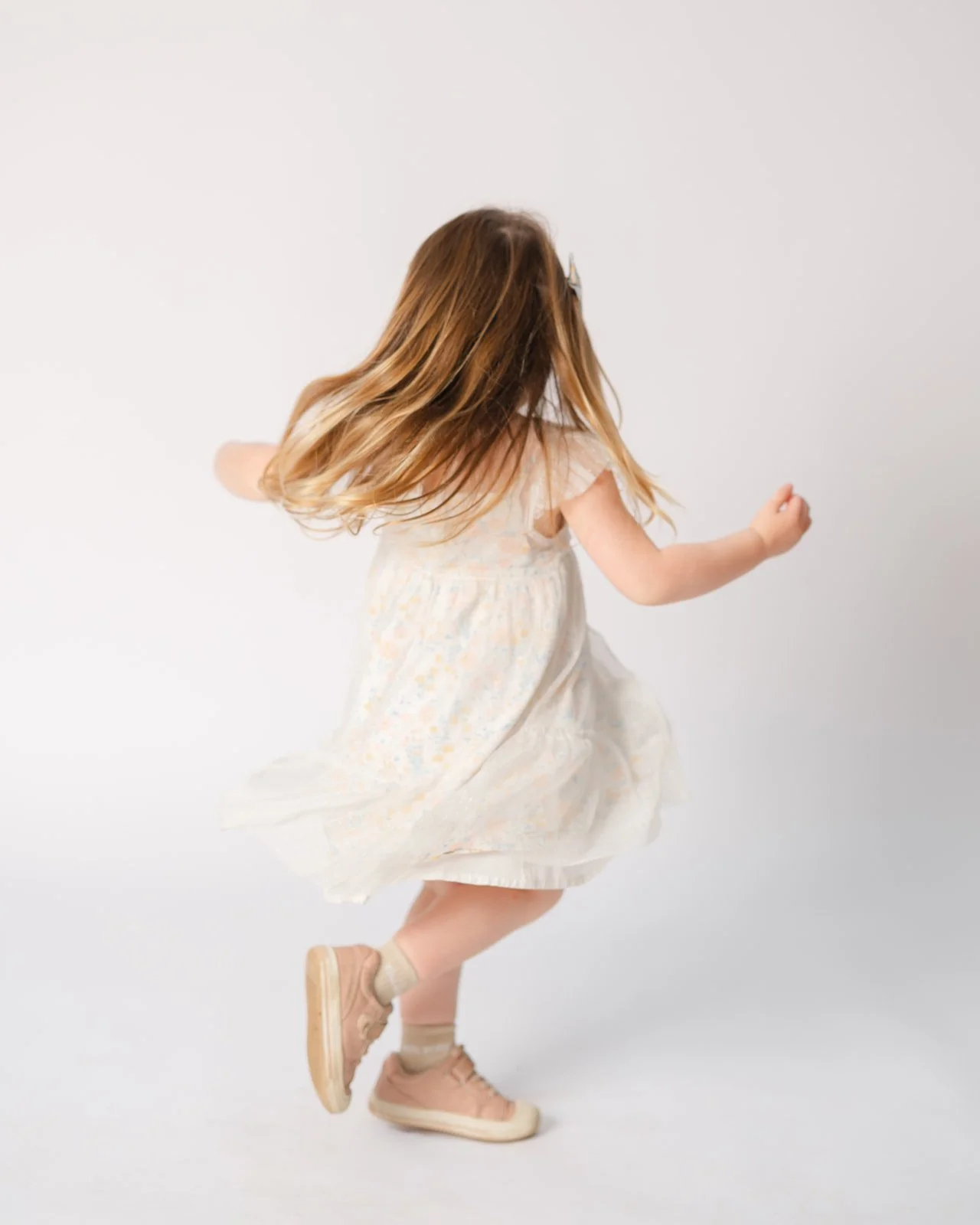A young girl with long, wavy, blonde hair is captured from behind, twirling in a white, flowy dress with a delicate, pastel floral pattern. She is wearing beige sneakers and beige socks, and is spinning with her arms slightly raised against a plain w
