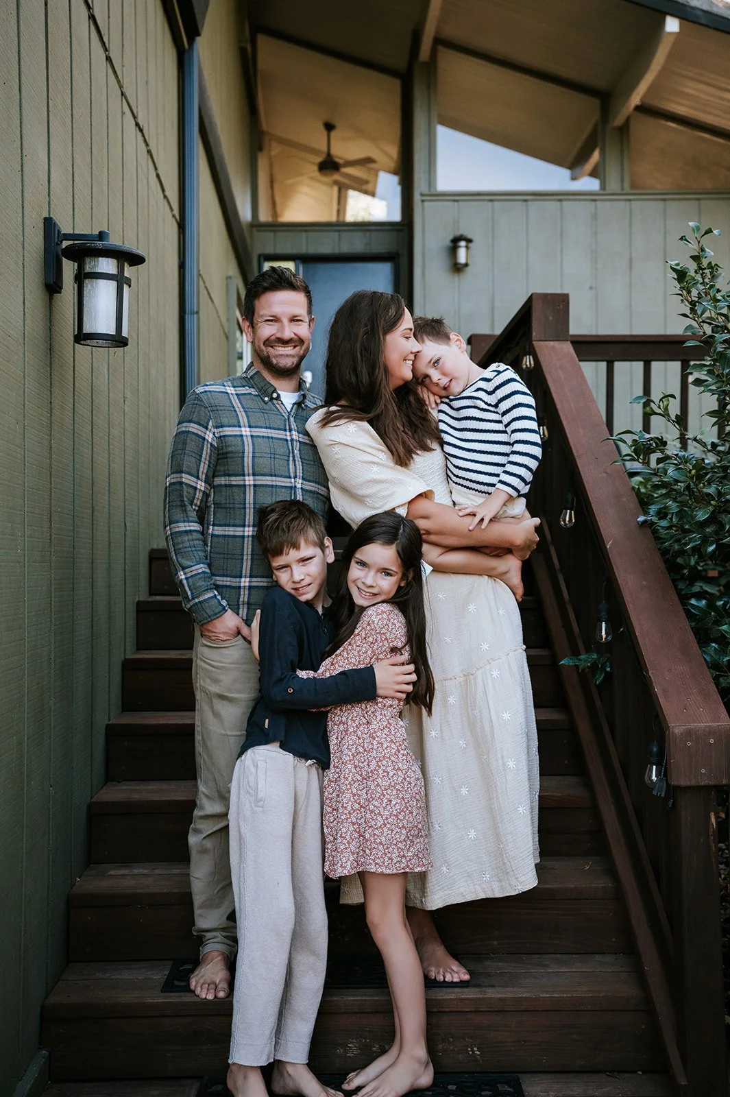 A family of six, including two men, one woman, and three children, standing on a wooden staircase outside a house. They are smiling and hugging, with the woman holding a young boy, and other children standing close with arms around each other.