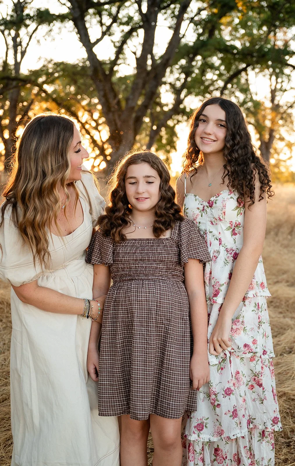 Three women and a girl standing outdoors during sunset, smiling and posing for a photo, with trees in the background.
