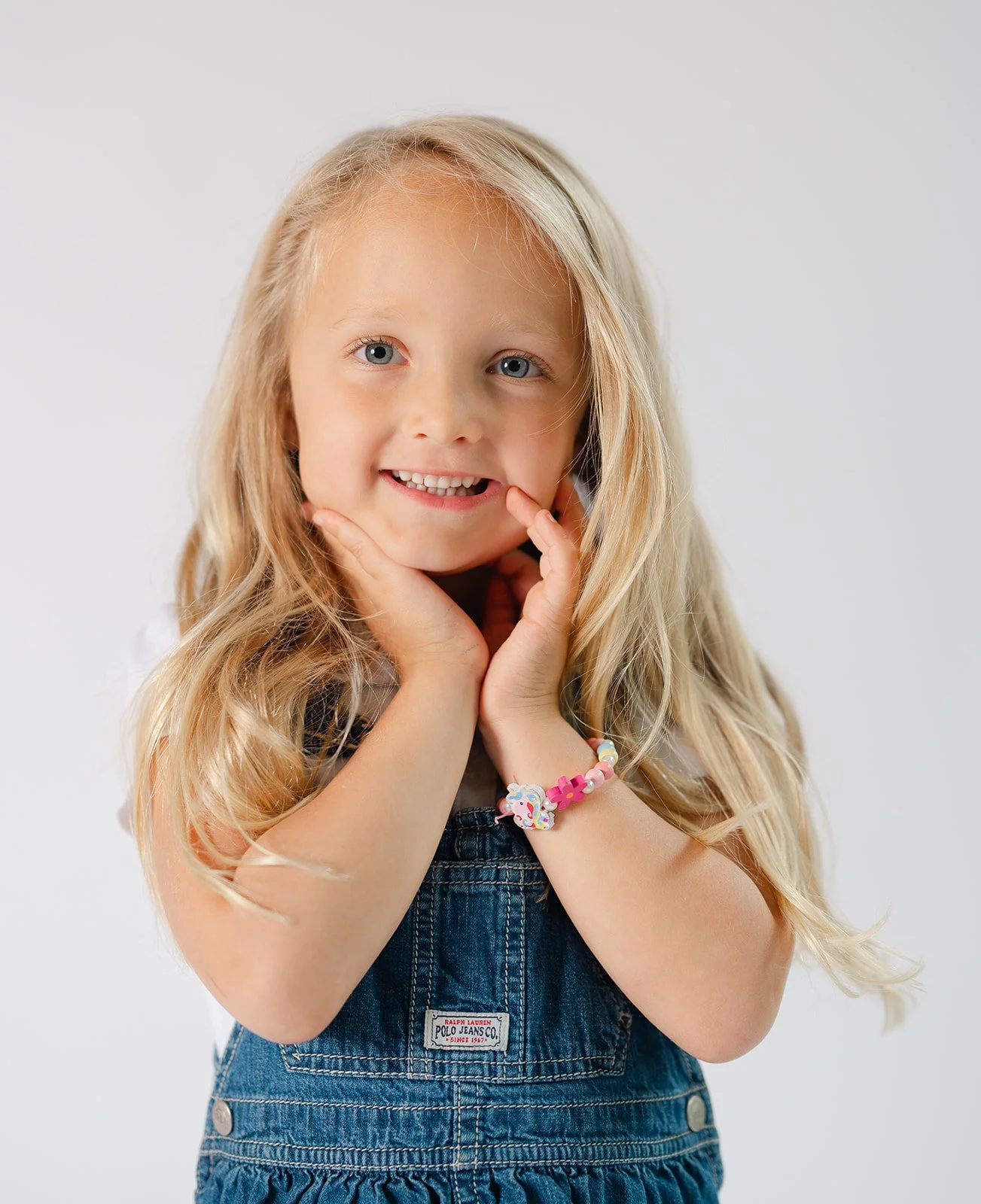 Smile of a young girl with long blonde hair, wearing a denim dress and a colorful bracelet, posing against a plain white background.