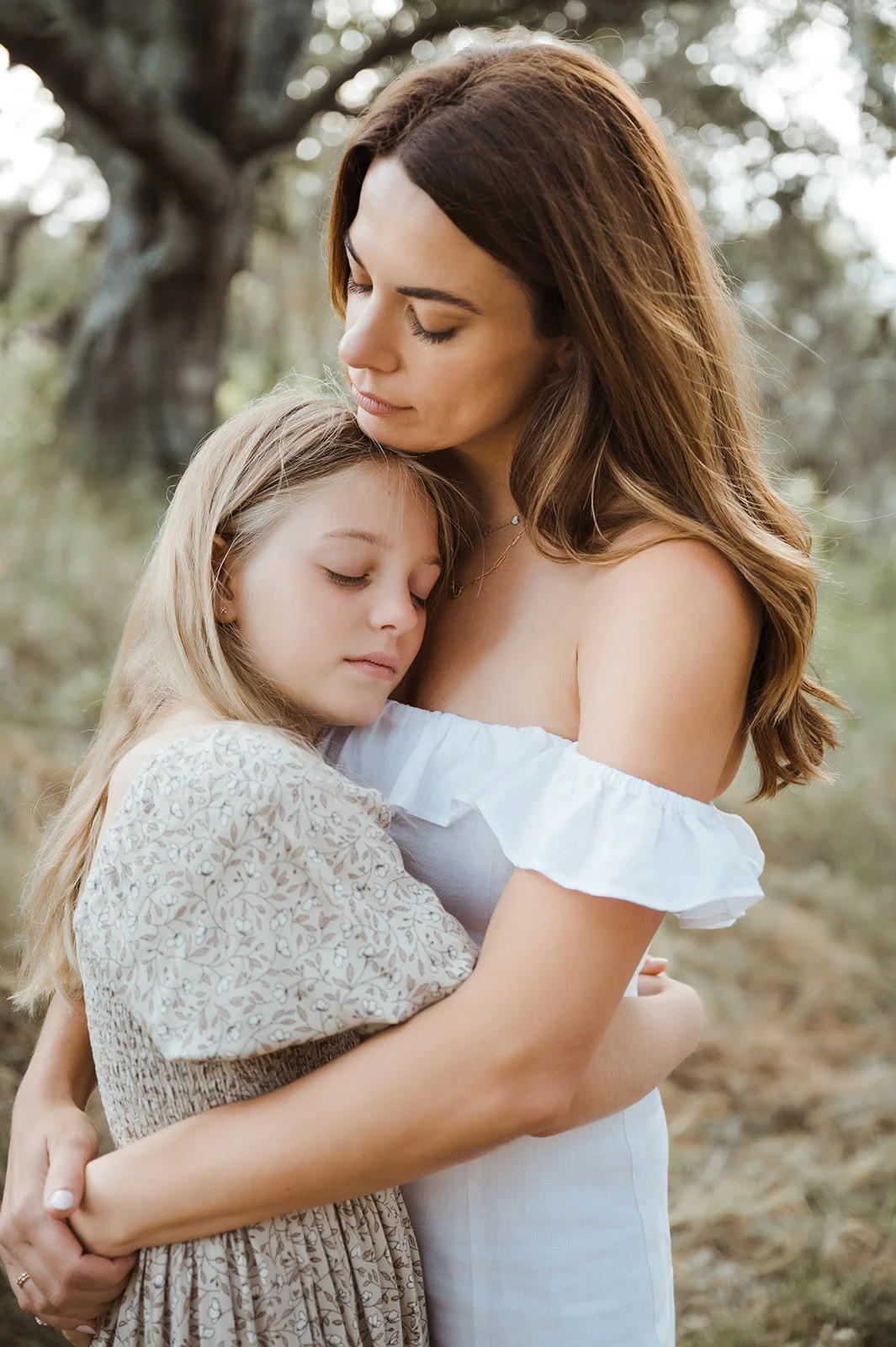 A woman with long brown hair hugging a young girl with blonde hair outdoors in a wooded area.